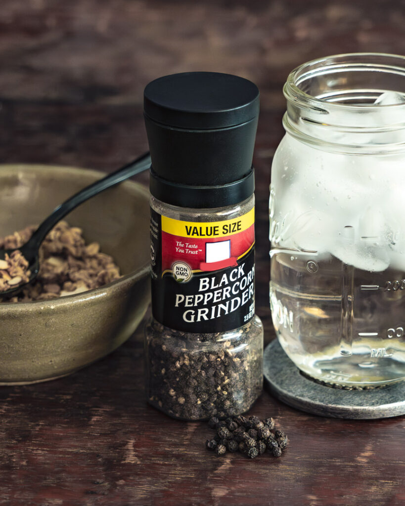 Black Pepper grinder and peppercorns, a glass of water, and a bowl of granola
