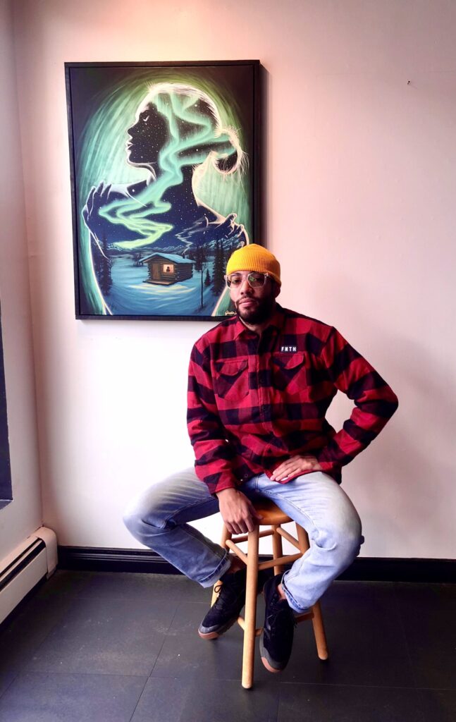 Artist Justin DeWolf seated beside his Alaska-themed cannabis artwork featuring northern lights and wilderness cabin, on display