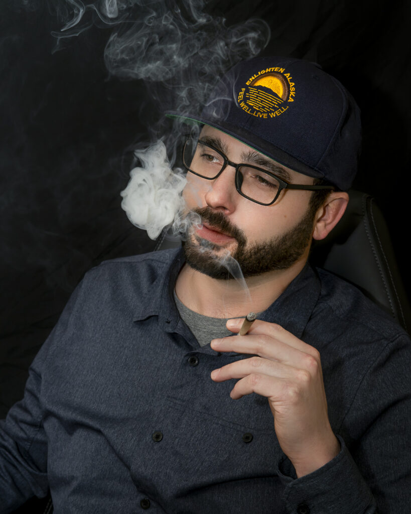 Enlighten Alaska CEO Evan Levinton smoking an Enlighten branded joint wearing an Enlighten Alaska branded hat against a dark background